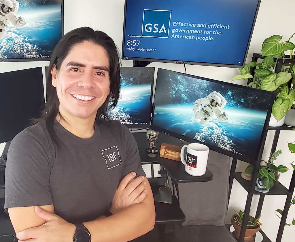 Edwin smiles at the camera crossing his arms. He has tan skin and long black hair. Behind him you can find his desk. There are four monitors on his desk. One is turned off, two have the image of an astronaut in space, and the fourth one has the GSA logo. On his desk there's also a mug with an 18F logo, a wooden ornament with the Puertorican flag, and a figurine of the Mandelorian. Next to his desk there's a shelf with green plants