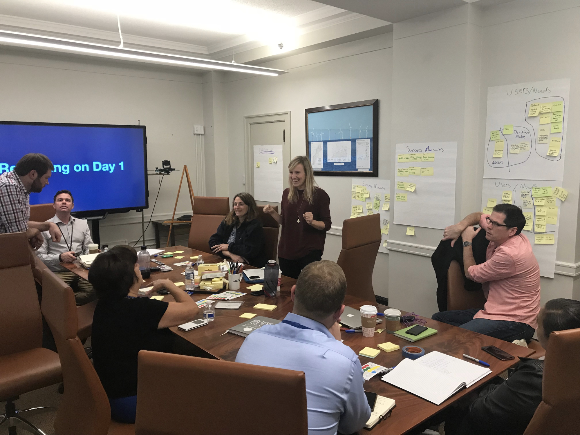 Product management workshop with 18F and ONRR. There is a group of people seating around a table with yellow stickie notes. On a wall there are long pieces of paper with yellow sticky notes on them