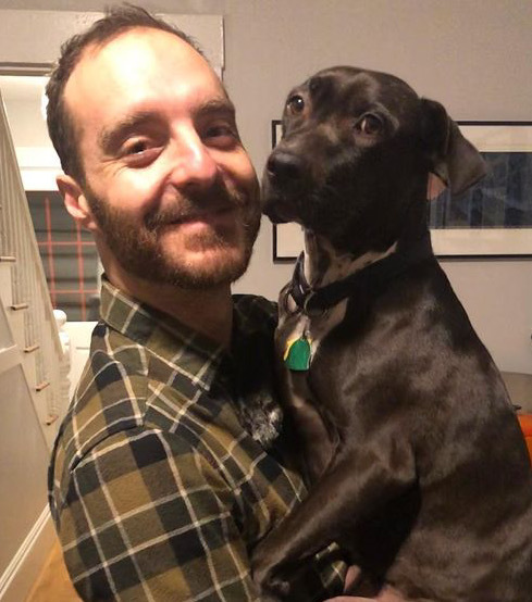 Mike Gintz (he/him) holding his really cute black dog smiling at the camera