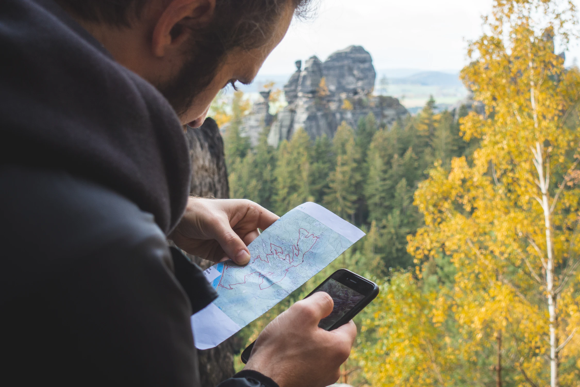 A person in the woods is looking at a paper map while also holding their smartphone.