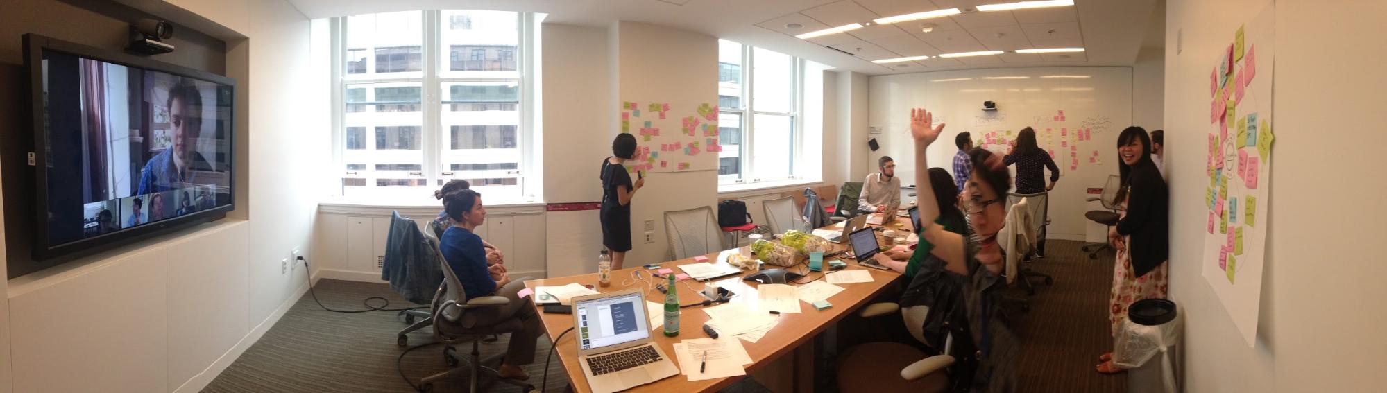 Image of a team in a meeting room. Various post its are stuck on the wall. To the left is a large screen tv with team members on a video call