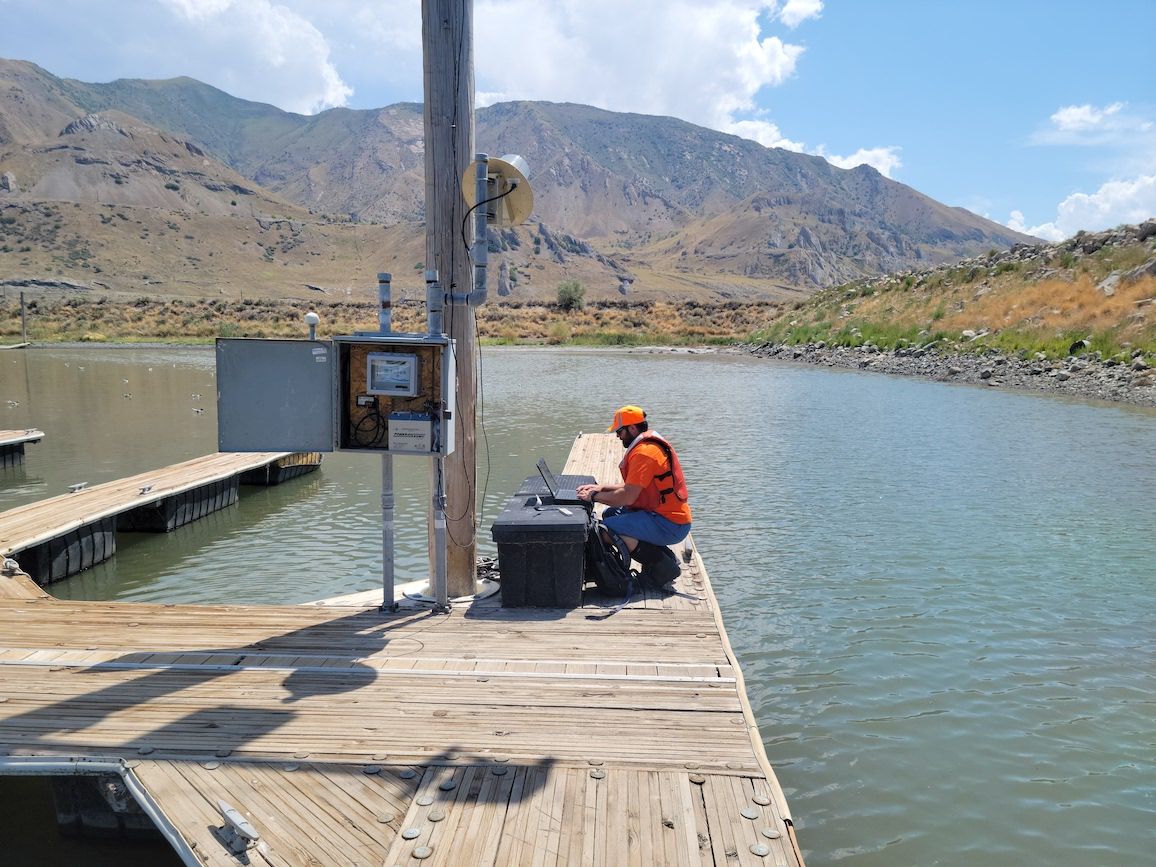 A worker in an orange hat and vest squats and types on a laptop, which is sitting on a plastic container on a pier in the Great Salt Lake. The Lakeside Mountains are in the background with partly cloudy skies.