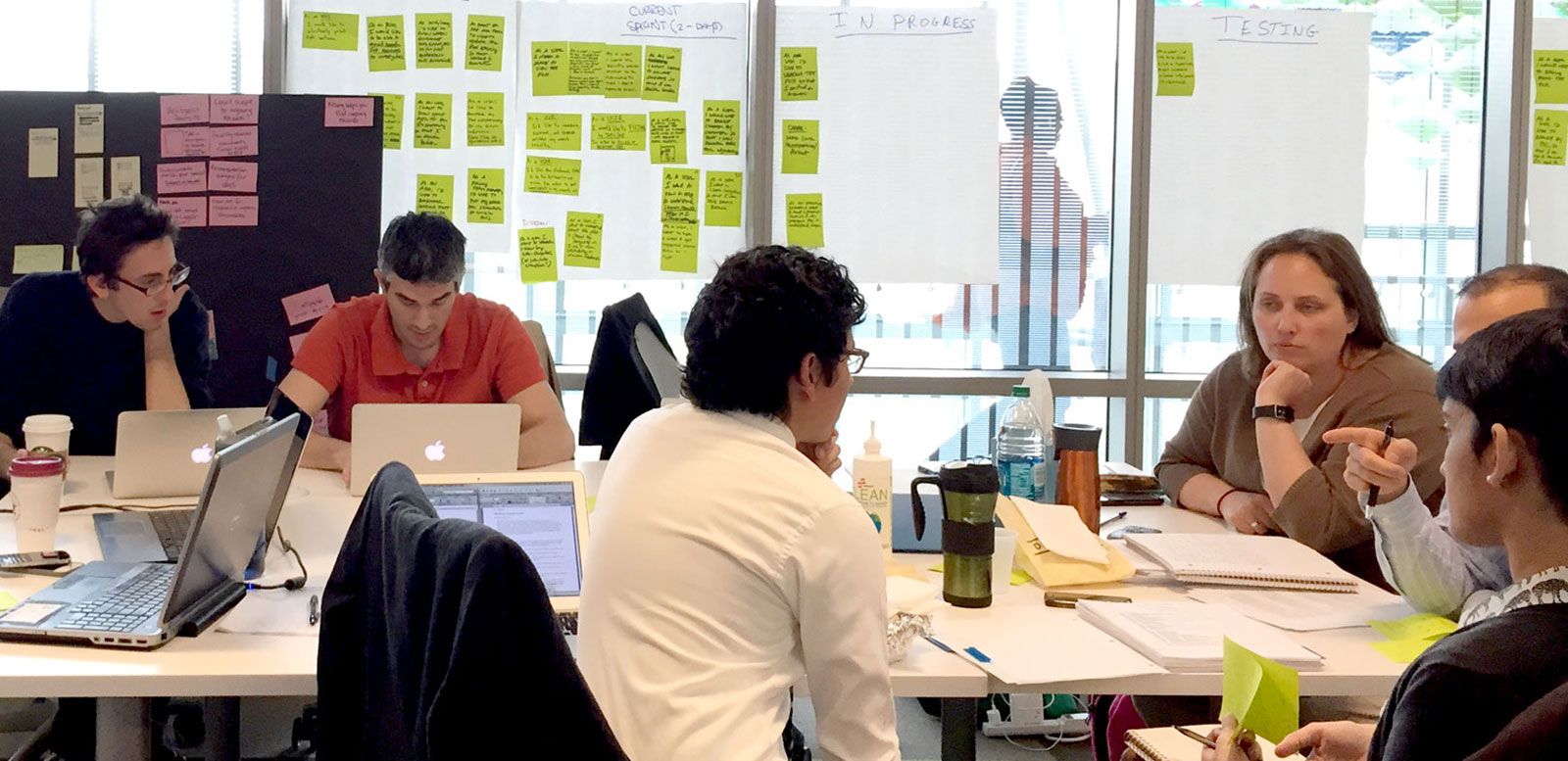 A group of civil servants collaborate on an 18F project. Two 18F colleagues work side-by-side across from product owners and investigators from the Department of Labor. The table is covered in laptops, notebooks, and coffee cups. Windows around the conference room have progress boards and sticky notes for the current 2-week sprint.