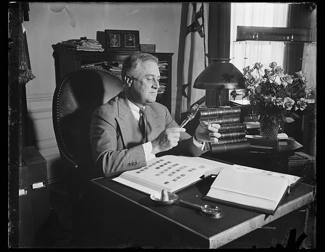 Franklin Roosevelt inspects stamps with a magnifying glass. He sits in a large office chair at a desk with leather books and roses. There's a short bookshelf behind him covered with periodicals.